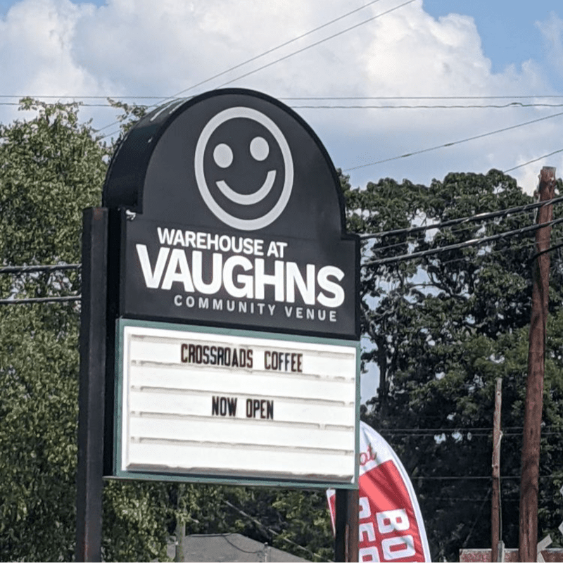 A black sign for the Warehouse at Vaughns Community Venue with a smiley face and a "Now Open" announcement for Crossroads Coffee stands against a backdrop of green trees and a cloudy blue sky.