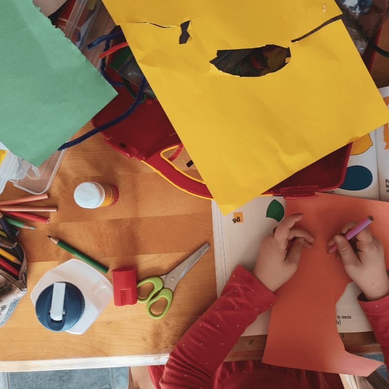 A child drawing on orange paper at a wooden table with craft supplies