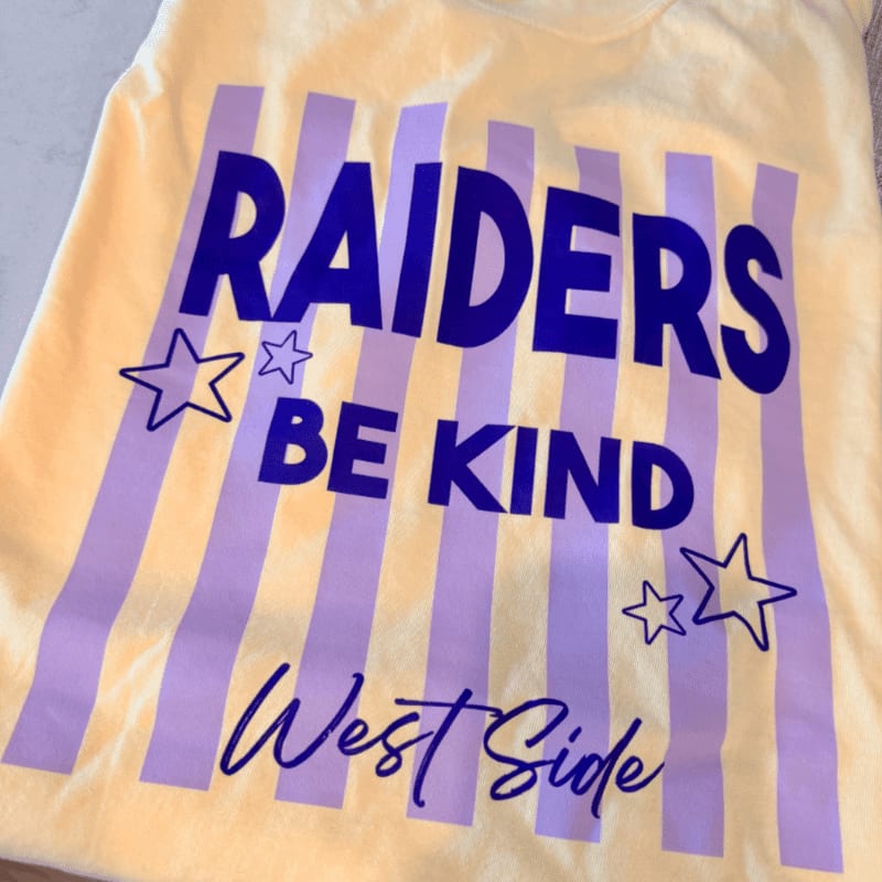 A yellow t-shirt features purple stripes and the text "RAIDERS BE KIND West Side" with star accents, set against a plain light background.