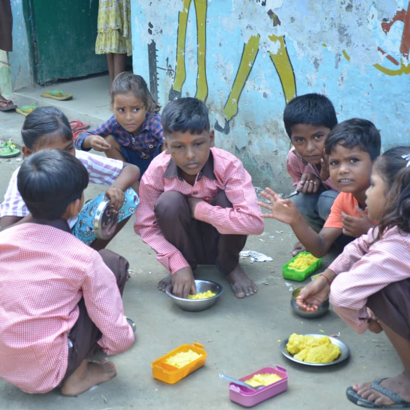 A group of young children in checkered uniforms sit on the ground eating food from bowls and containers against a weathered blue wall.