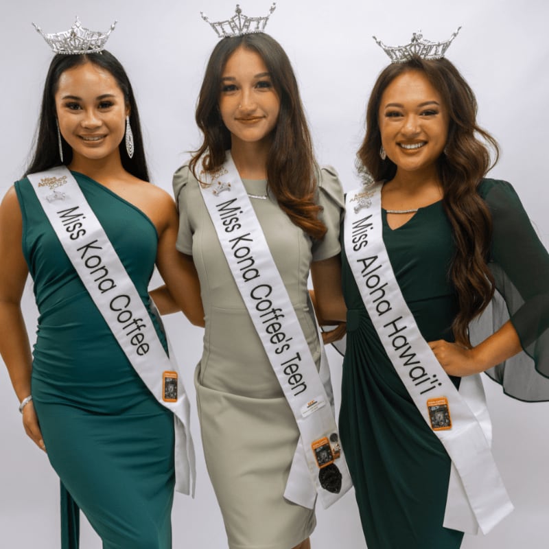 Three women wearing crowns and sashes stand side-by-side against a plain white background.