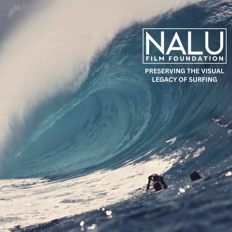 Two surfers float in the foreground beneath a massive, curling blue wave that dominates the background.