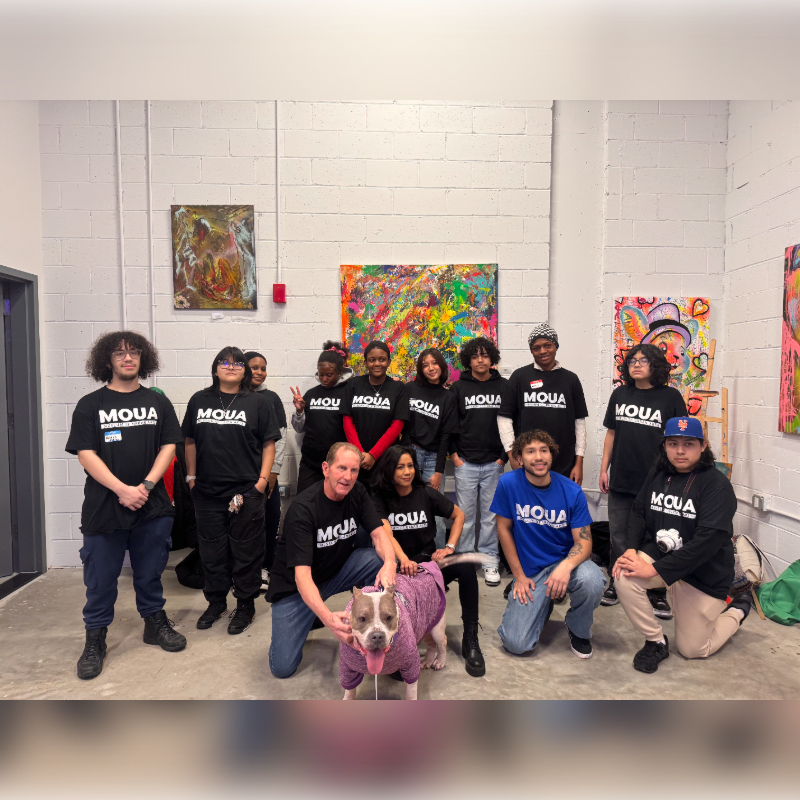 A group of people wearing matching "MOUA" t-shirts pose with a dog in a studio featuring white brick walls and colorful paintings.