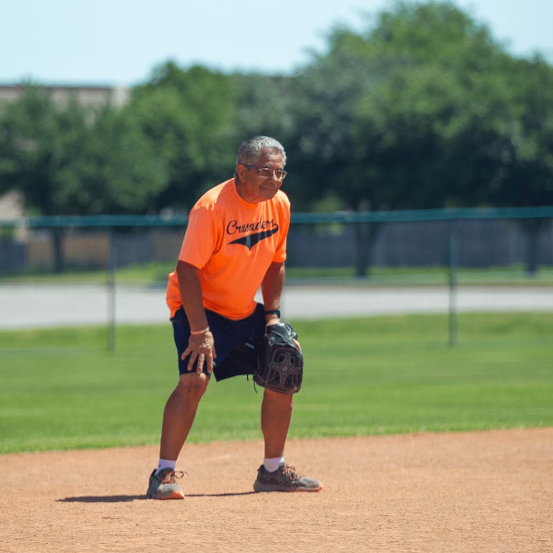 An older man in an orange shirt and baseball glove stands on a dirt field with green grass and trees in the background.