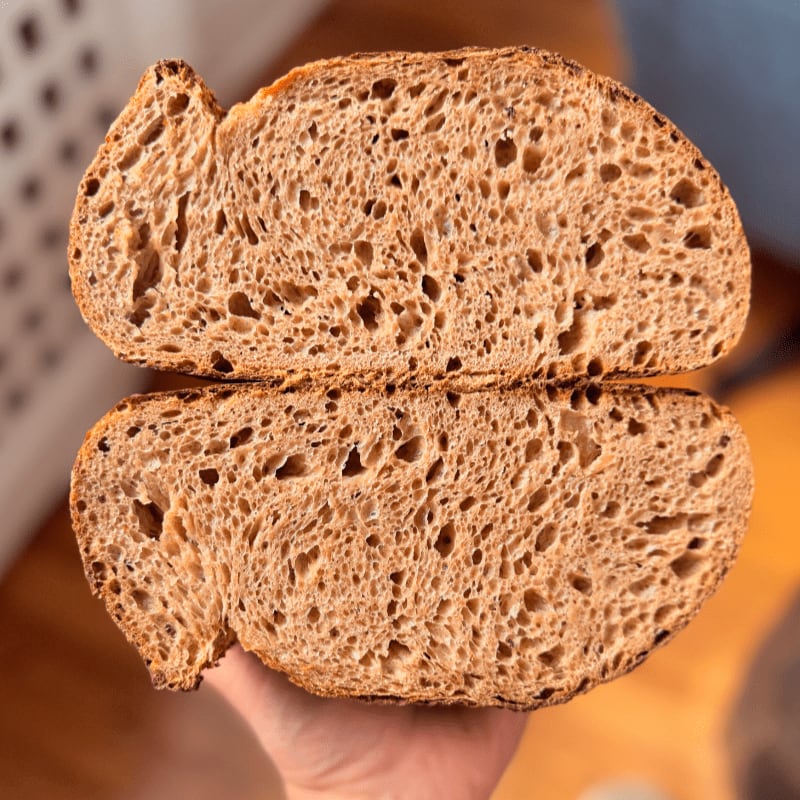 Two halves of a sliced sourdough loaf with an airy crumb are held up against a blurred indoor background.