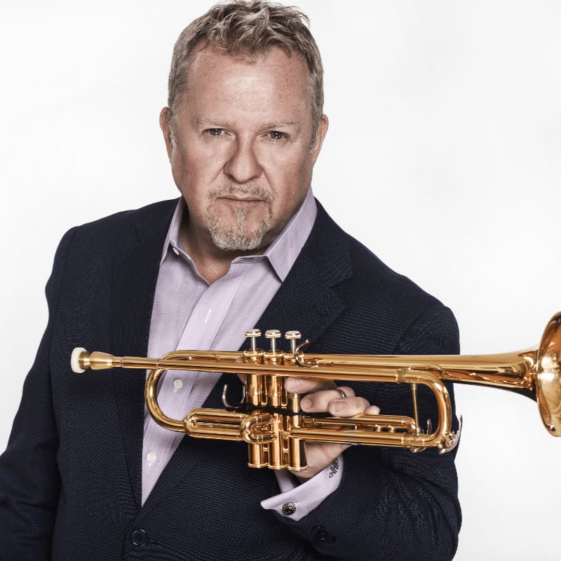 A man in a dark blazer and light shirt holds a gold trumpet against a plain white background.