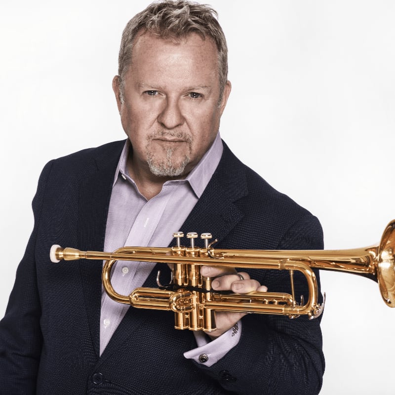 A man in a dark blazer and light shirt holds a gold trumpet against a plain white background.