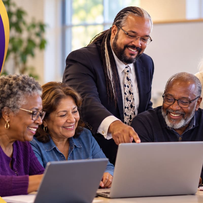 Three smiling older adults look at a laptop screen while a man in a suit points to the display in a bright, blurred office setting.