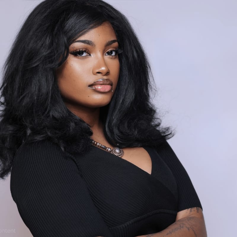A woman with voluminous black hair and a black ribbed top poses against a plain, light purple background.