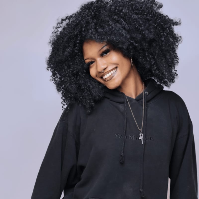 A smiling woman with voluminous curly black hair wearing a black hoodie stands against a plain light grey background.