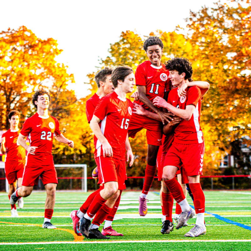 Soccer players in red uniforms celebrate on a green field against a backdrop of vibrant autumn trees.