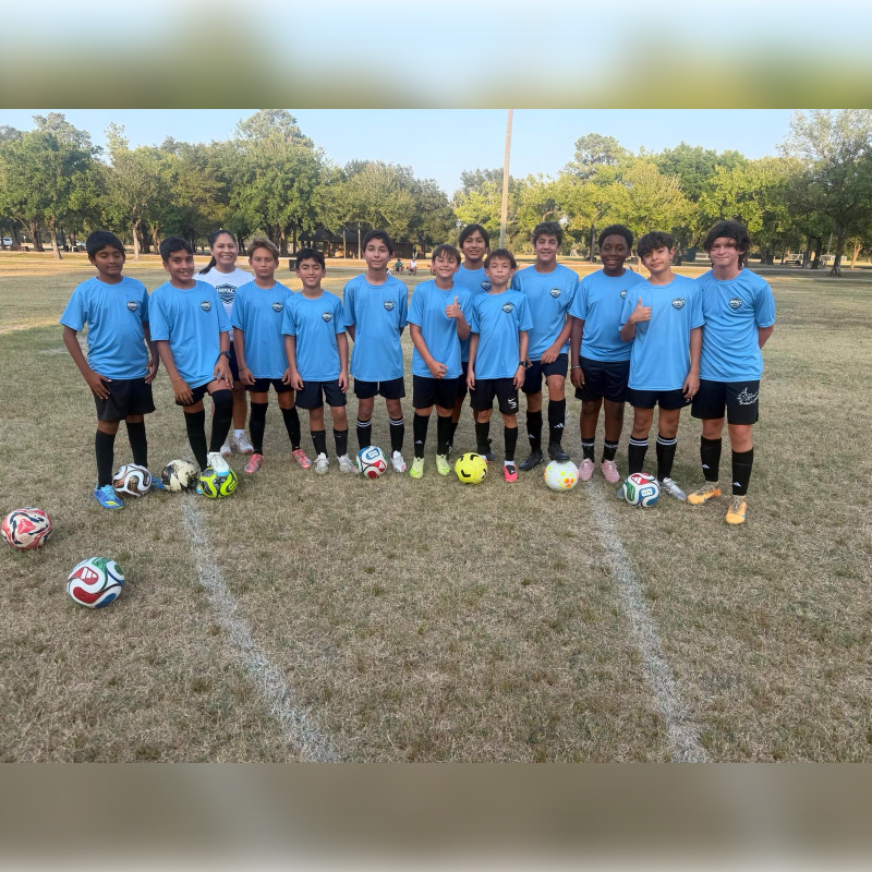 A youth soccer team in light blue jerseys stands together on a grassy field with soccer balls in the foreground and lush green trees in the background.