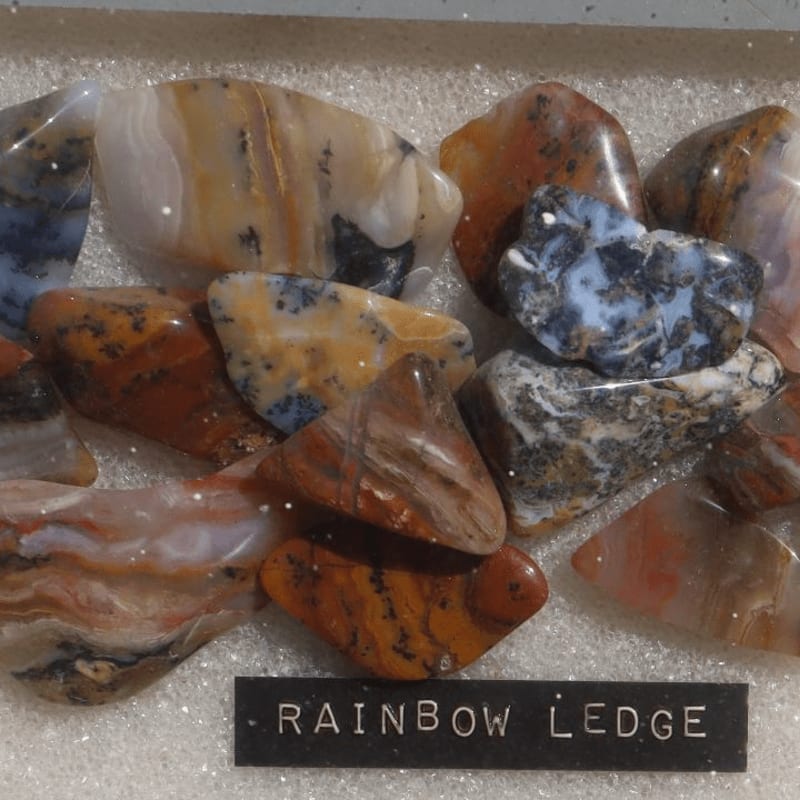 A collection of polished, multicolored stones sits atop a white shimmering surface with a black label reading "RAINBOW LEDGE" in the foreground.