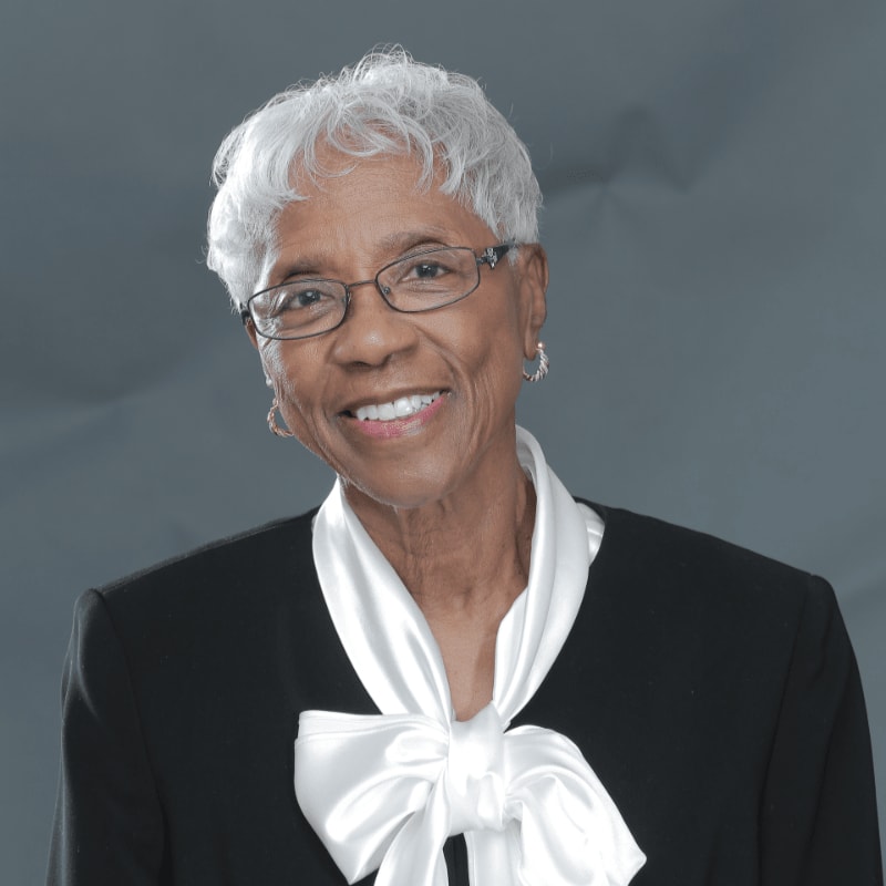 A smiling woman with short white hair and glasses wearing a black blazer and white bow tie stands against a plain grey background.