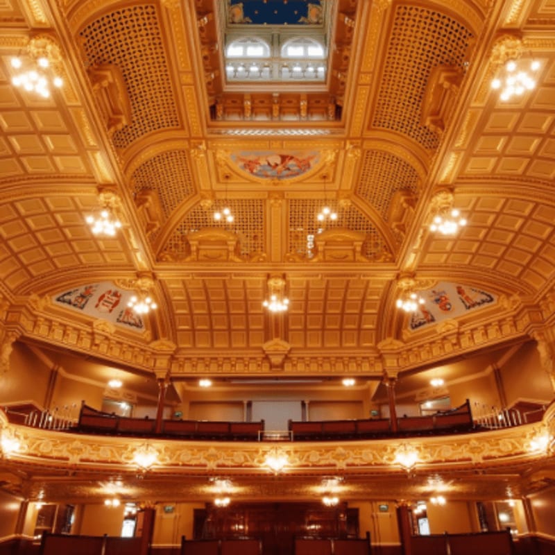 An ornate, gold-toned ceiling with intricate carvings and paintings looms over a tiered wooden balcony and seating area.