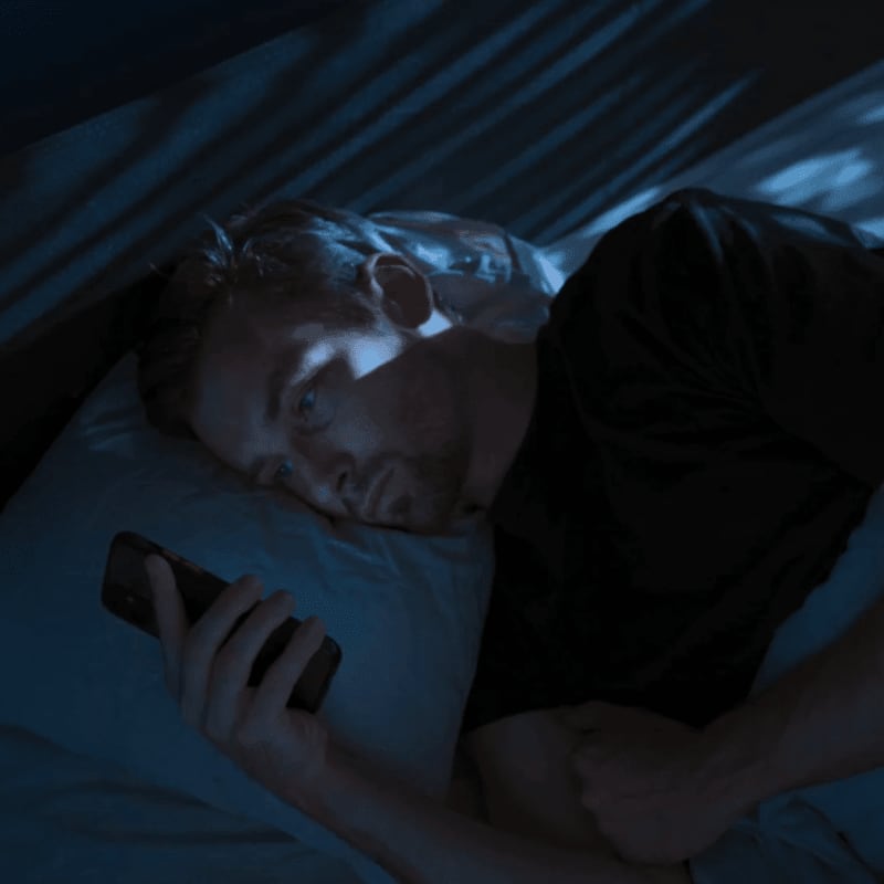 A man lies in bed in a dark room, illuminated by the blue light of a smartphone he is holding in the foreground.