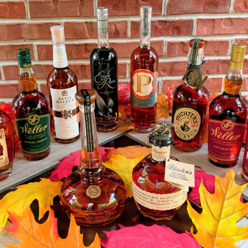 Various bottles of bourbon are arranged on a surface with autumn leaves in the foreground against a red brick wall background.