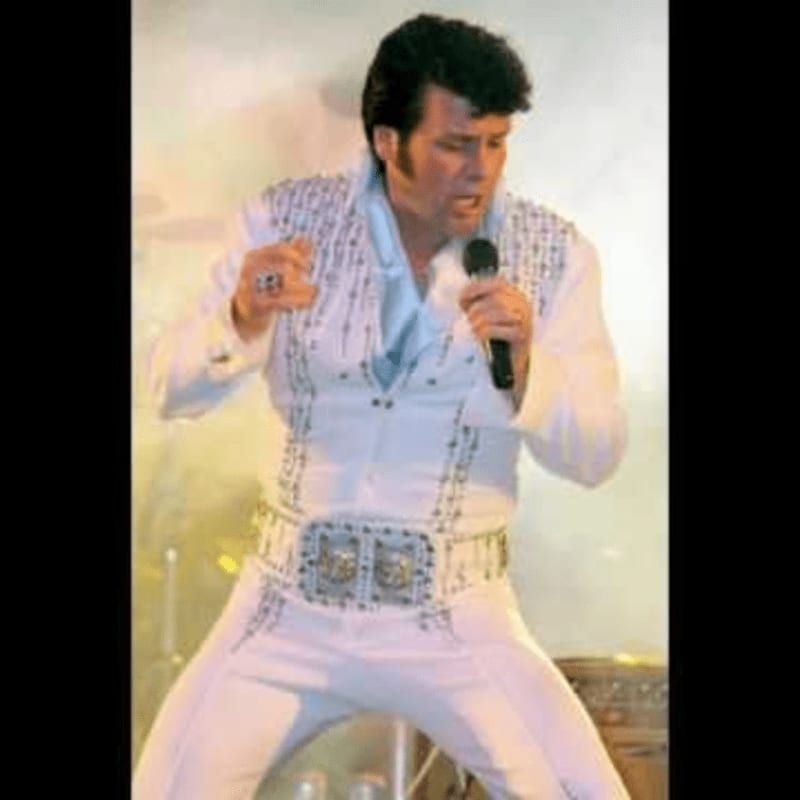 A man in a white sequined jumpsuit sings into a microphone against a plain, light-colored background.