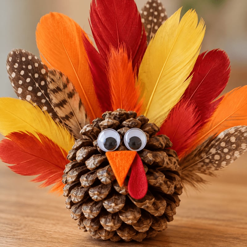 A pinecone turkey with googly eyes and colorful feathers sits on a wooden surface against a blurred background.