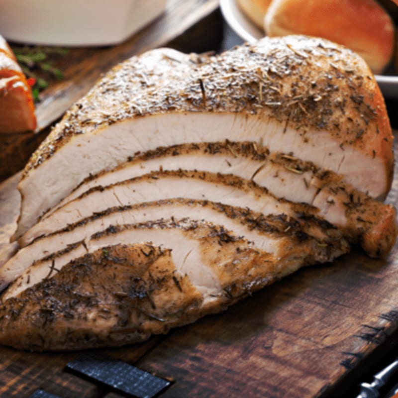 Sliced herb-roasted turkey breast rests on a wooden cutting board with rolls and a white dish in the background.