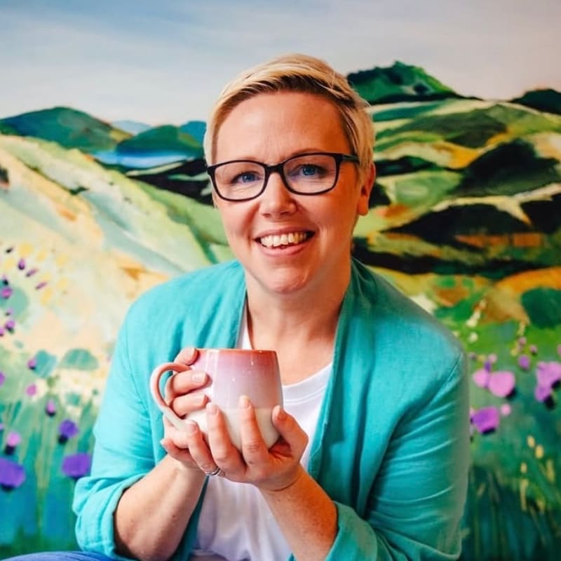 A smiling woman in a turquoise blazer holds a pink and white mug in front of a colorful landscape painting.