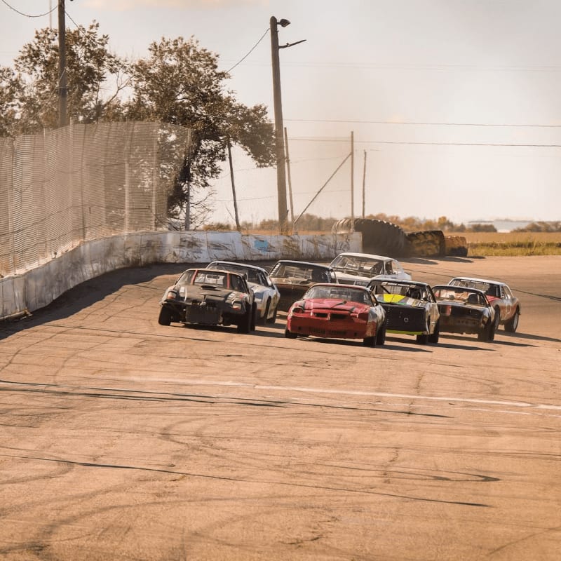 Several beat-up race cars compete on a paved track bordered by a white wall and chain-link fence under a clear sky.