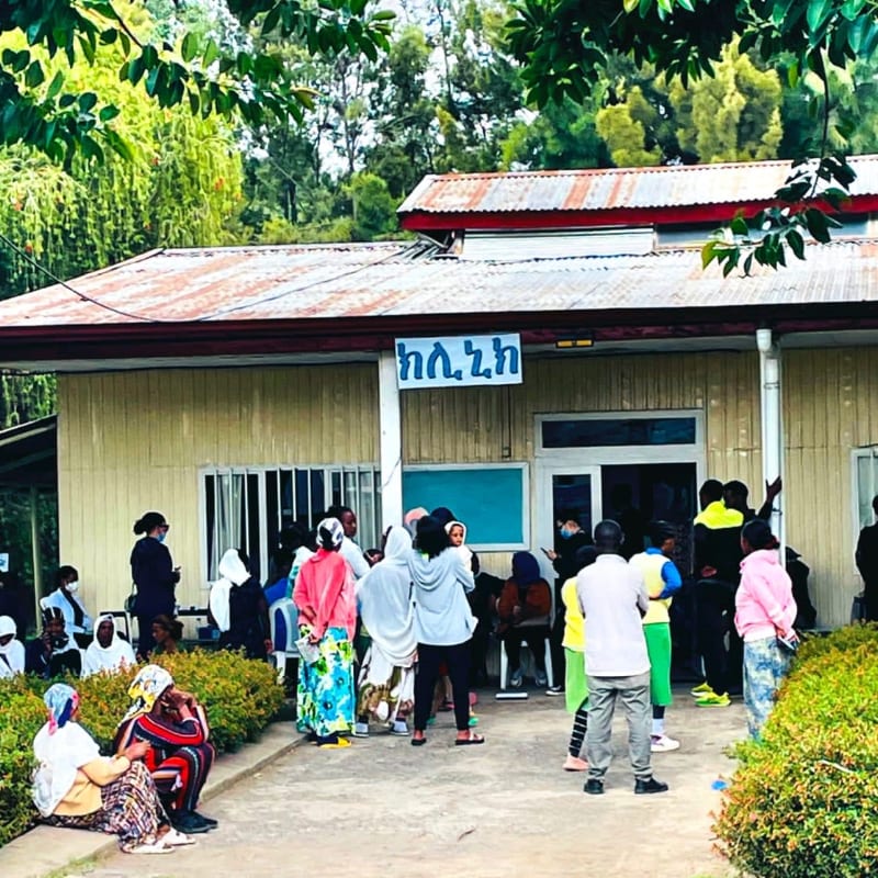 A group of people gather in front of a small building with a corrugated metal roof, surrounded by lush green trees and shrubbery.