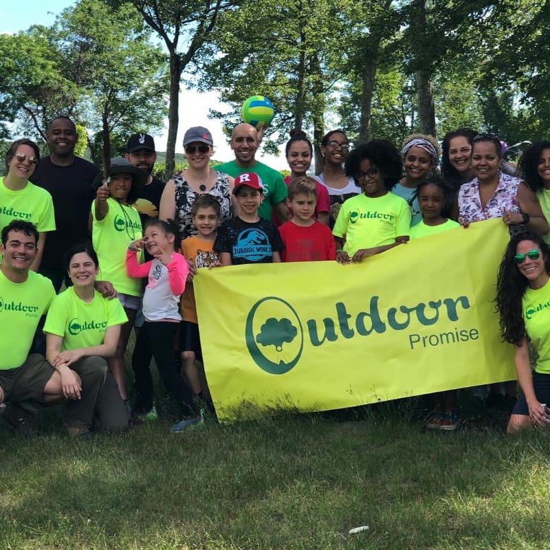 A diverse group of adults and children pose with a yellow "Outdoor Promise" banner in a grassy field backed by lush green trees.