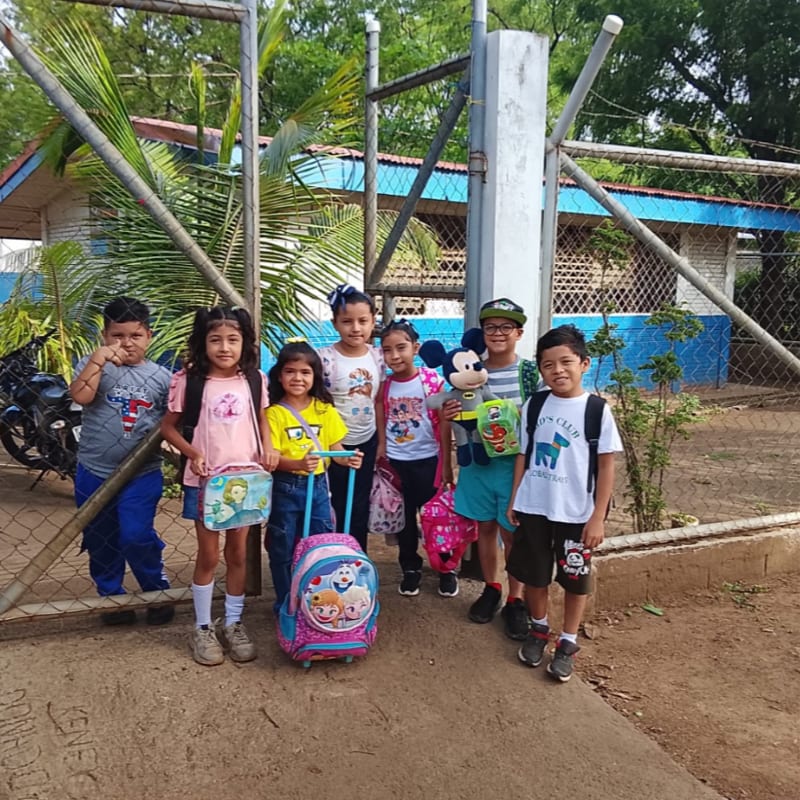 Six young children stand together in the foreground, while a chain-link fence and a blue-and-white building sit in the background.