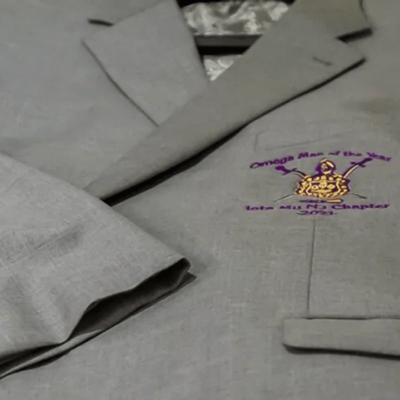 A grey linen garment with a purple embroidered logo sits atop a matching grey fabric background.