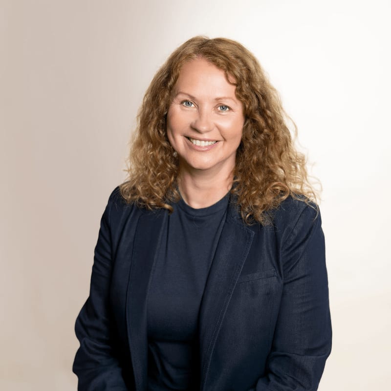 A smiling woman with curly reddish-blonde hair wearing a navy blue blazer stands against a plain, light-colored background.