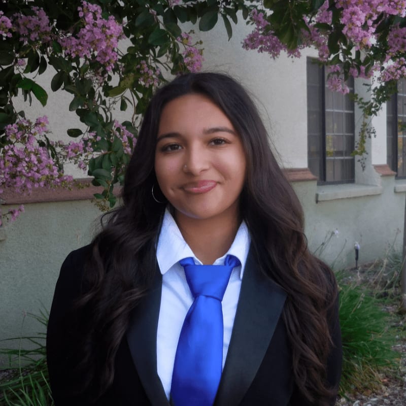 A young woman wearing a black blazer, white shirt, and blue tie stands in the foreground against a background of a white building and pink flowering bushes.