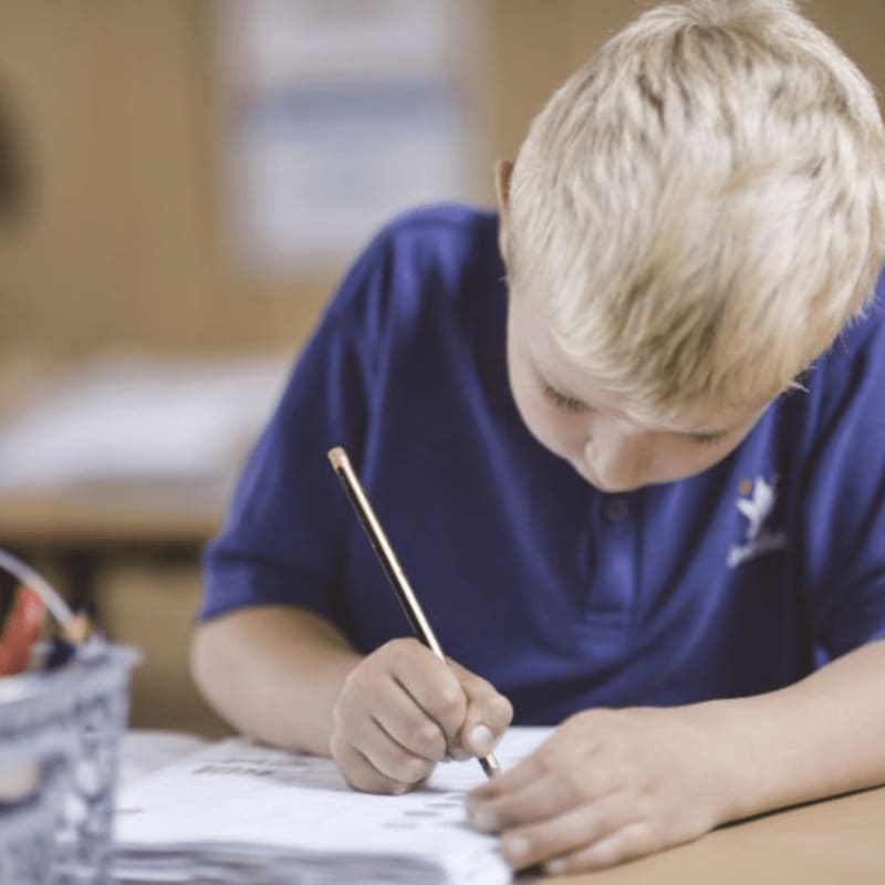 A young boy in a blue shirt writes on a piece of paper in the foreground, with a blurred classroom setting in the background.