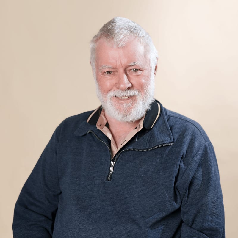 An older man with a white beard and a blue quarter-zip sweater stands against a plain, light-beige background.