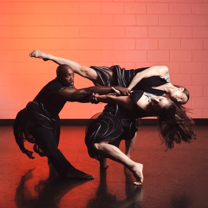 Three dancers perform a dramatic, intertwined pose in the foreground against a red-lit brick wall background.
