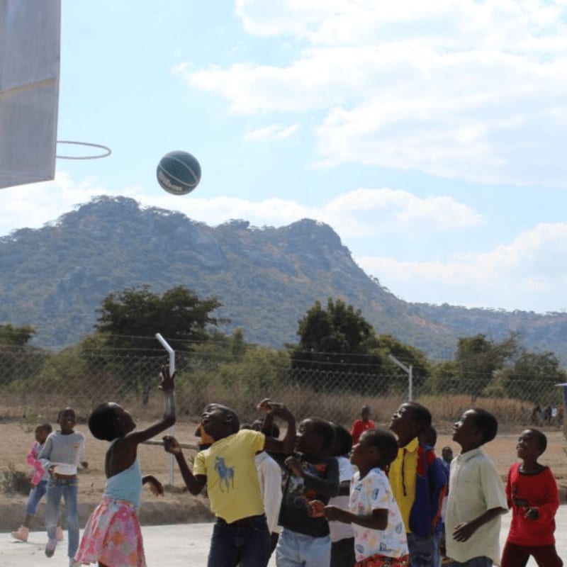 A group of children look up at a basketball in mid-air on an outdoor court with mountains and a blue sky in the background.