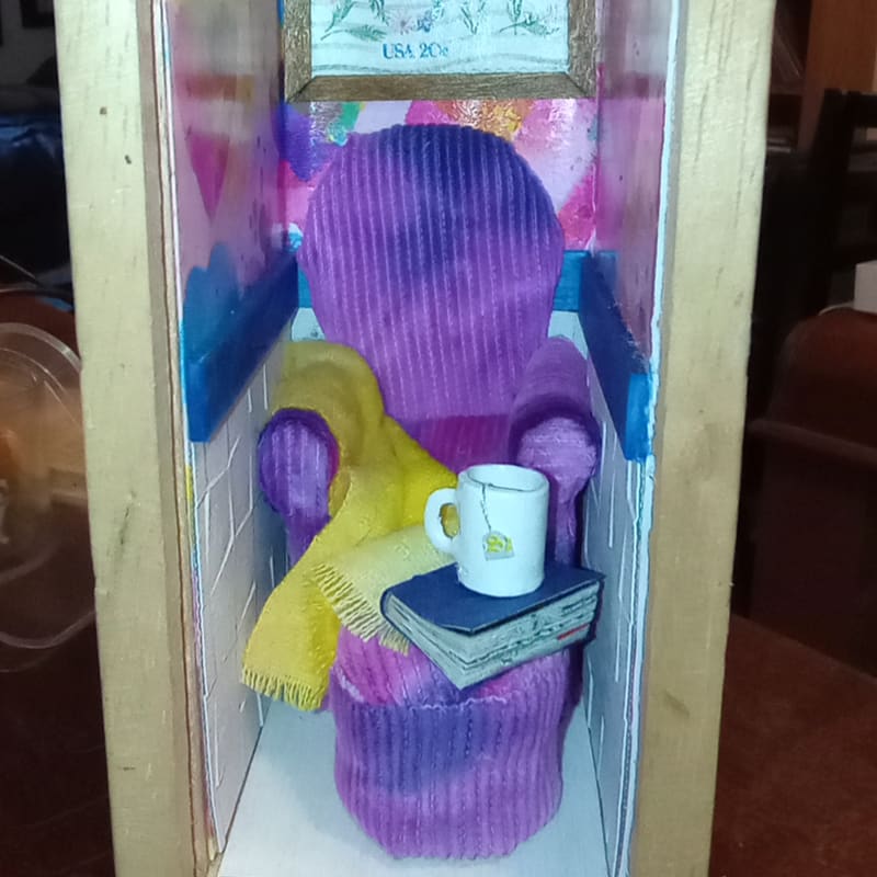 A miniature purple armchair with a yellow blanket, a book, and a white mug sits inside a wooden box against a colorful, artistic background.