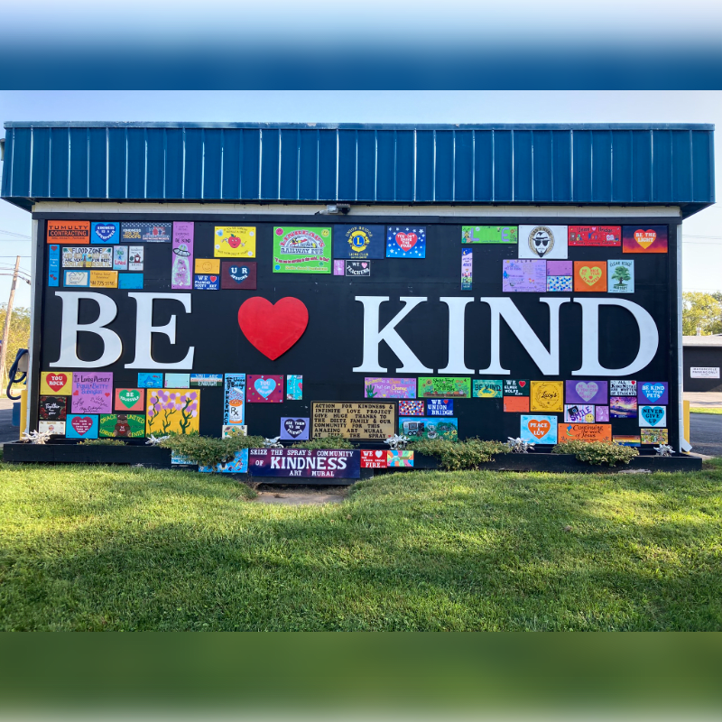 A large "BE KIND" mural with a red heart and colorful tiles covers a building wall behind a green grassy lawn.