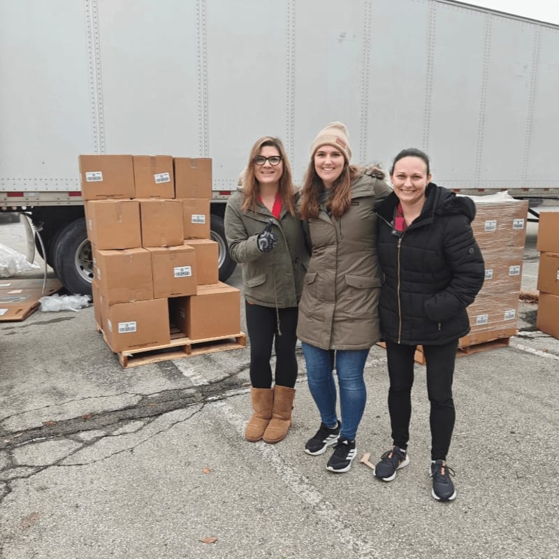 Three women stand smiling in the foreground next to stacks of cardboard boxes, with a large white semi-trailer in the background.