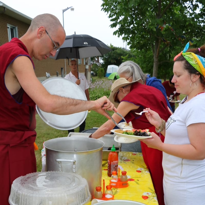 A man in maroon robes serves food from a large pot to people at an outdoor gathering with a house and trees in the background.