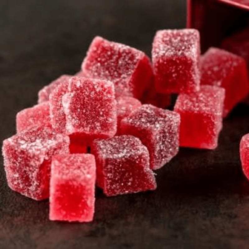 A pile of sugar-coated red gummy candies sits on a dark, textured surface with a red container in the background.