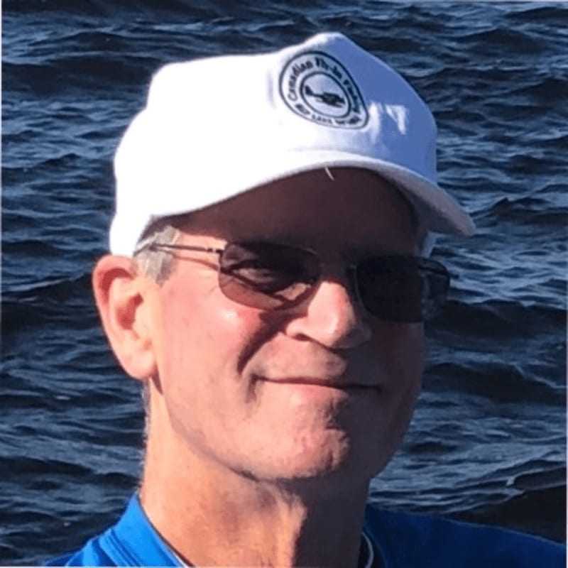 A man wearing a white cap and sunglasses stands in the foreground against a background of dark blue water.