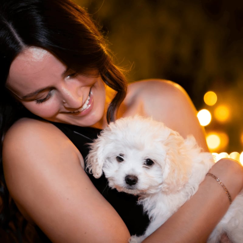 A smiling woman holds a small white puppy in the foreground against a dark, blurred background with golden bokeh lights.