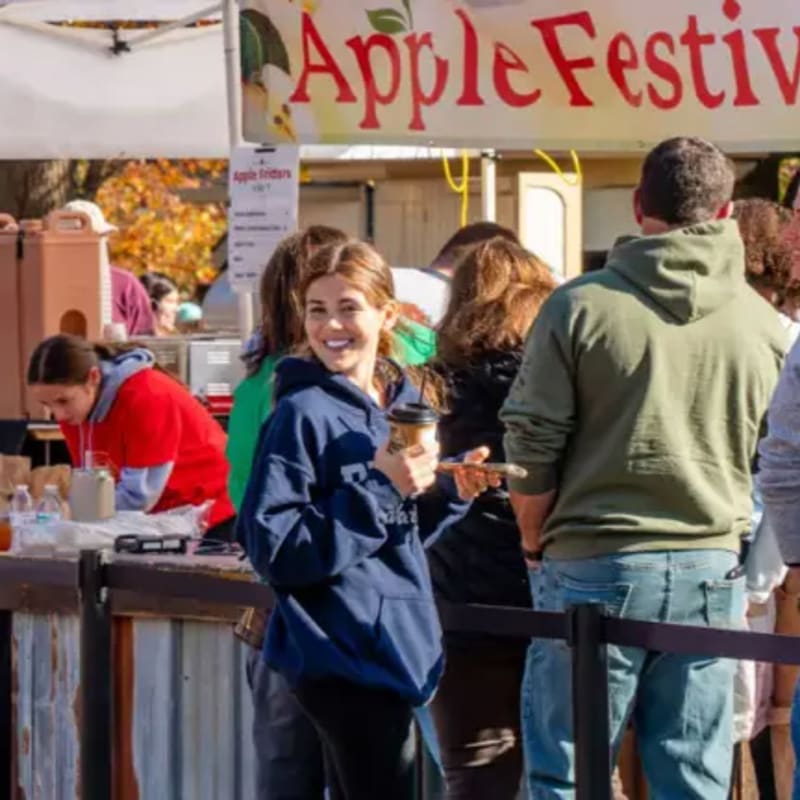 A smiling woman in a blue hoodie holds a coffee cup in the foreground, while other people stand before an "Apple Festival" banner in the background.