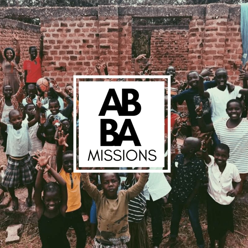 A group of smiling children gathers in the foreground in front of a rustic brick wall background, with a white "ABBA MISSIONS" logo centered over the scene.