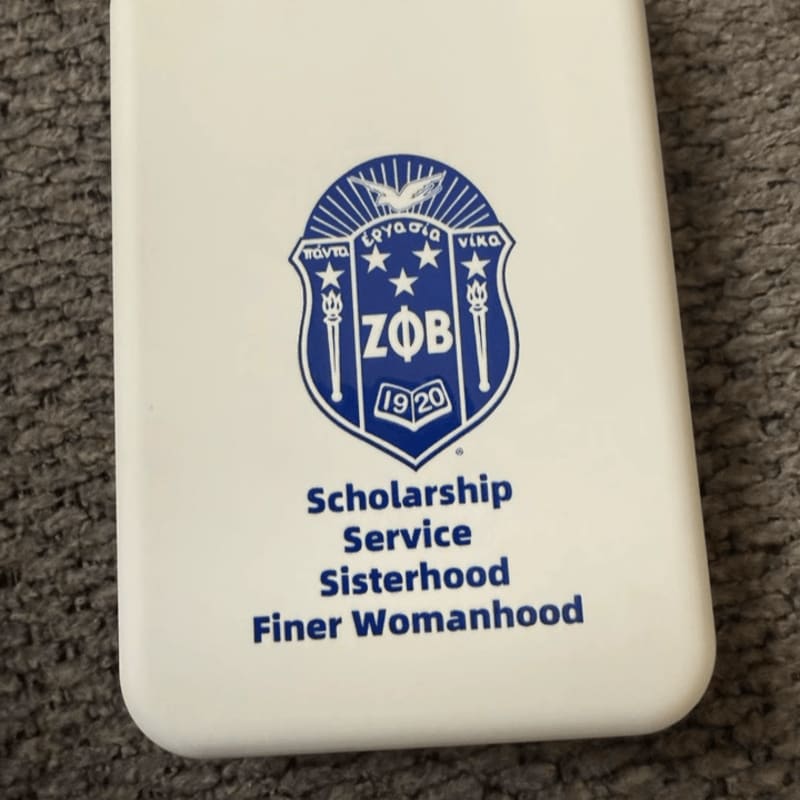 A white rectangular object featuring a blue Zeta Phi Beta sorority crest and motto rests on a grey textured carpet.