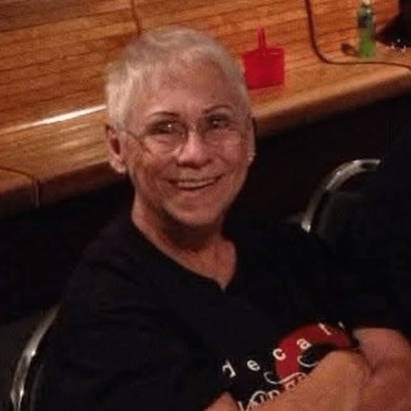 A smiling woman with short grey hair and glasses wearing a black t-shirt sits in front of a wooden ledge with a red cup and a water bottle.