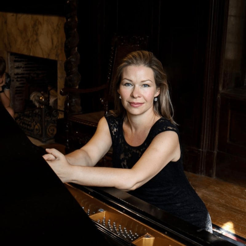 A woman in a black dress leans against a piano in a dimly lit room with a fireplace and ornate furniture in the background.
