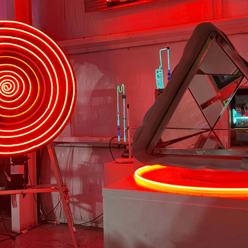 A glowing red neon spiral stands in the foreground beside a mirrored geometric structure, with blue neon tubes visible in the background.