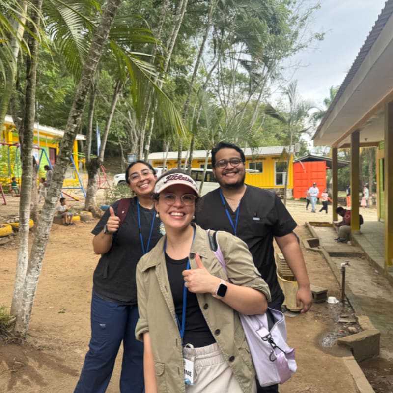 Three smiling people pose for a photo in the foreground, with tropical trees and yellow buildings in the background.